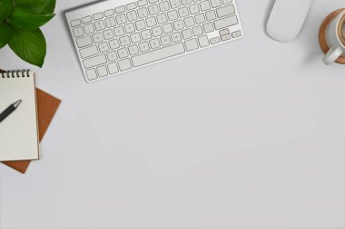 Simple workspace with blank notepads, coffee cup, wireless keyboard and houseplant on white table.