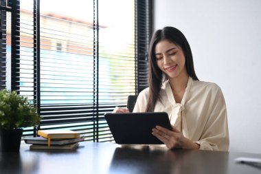 Satisfied working woman sitting in contemporary office and using digital tablet.