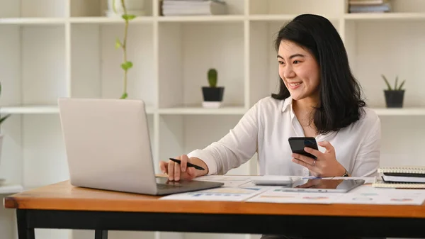 Pretty Asian Female Employee Holding Mobile Phone Using Laptop Wooden — Zdjęcie stockowe