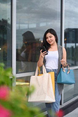 Portrait of young woman with shopping bags using smart phone and walking in the city street.