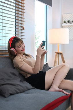Joyful young woman wearing wireless headphone using mobile phone on couch in comfortable home.