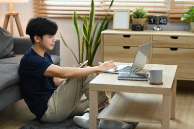 Asian man working online from home with laptop computer, sitting in bright living room.