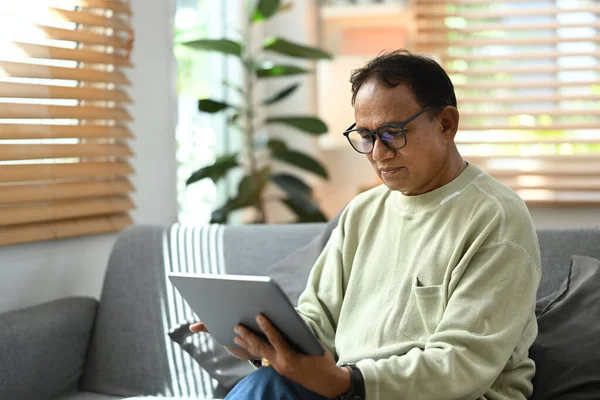 Calm mature man wearing eyeglasses reading online news on digital tablet while resting on couch in bright living room.