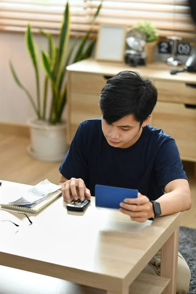 Focused man using calculator for calculating money bank loan rent payments, managing expenses finances in bright living room.