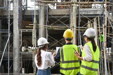 Rear view of female investor and civil engineer in hardhats and reflective jackets standing front of commercial building construction site.