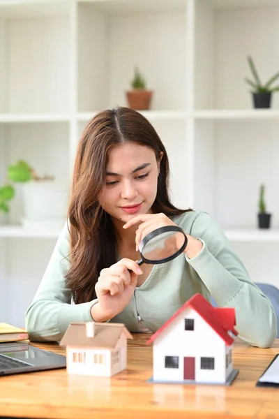 Young woman appraiser with magnifying glass looking at house models. House search and land valuation concept.