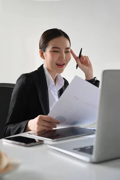 Happy asian woman in business suit working with financial document and laptop at bright office.
