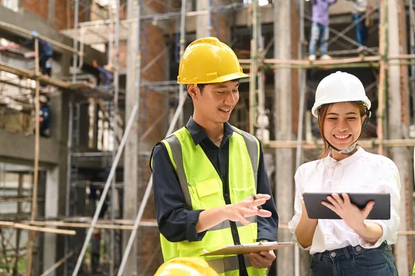 Smiling male supervisor and female investor wearing hardhats and reflective jackets inspecting commercial building construction site.