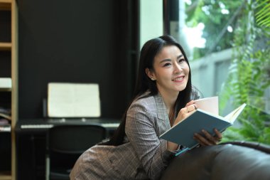 Elegant asian businesswoman resting on couch in bright modern office and reading book.