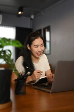 Charming female freelancer reading online news or checking email on laptop, sitting in her home office.