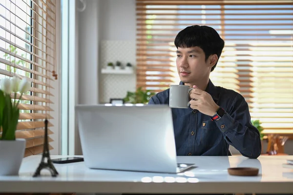 Thoughtful young businessman drinking coffee coffee and looking through window , spending free time in morning at his home office.