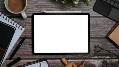 Top view digital tablet with blank display, surrounded by coffee cup, glasses and supplies on wooden office desk.