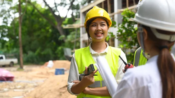 Smiling female engineer supervisor and investor wearing safety helmets discussing the construction process together while visiting a new building.
