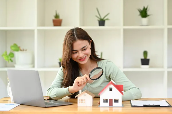 Smiling young female appraiser looking at house model through magnifying glass. Concept of real estate appraisal, land valuation, house search.