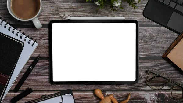 Top view digital tablet with blank display, surrounded by coffee cup, glasses and supplies on wooden office desk.