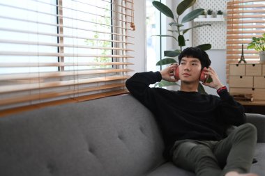 Satisfied man wearing warm sweater listening to music on headphones, spending leisure time in cozy autumn weekend at home.