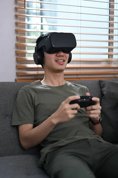 Excited man holding joystick and playing video game with virtual reality glasses on couch.
