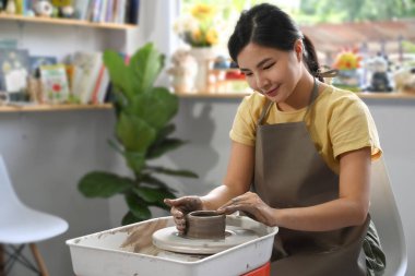 Concentrated asian female potter sculpting clay dishes on the pottery wheel in creative studio workshop.