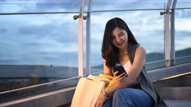 Happy young woman using smart phone while sitting on stairs at rooftop with background of cityscape at sunset.