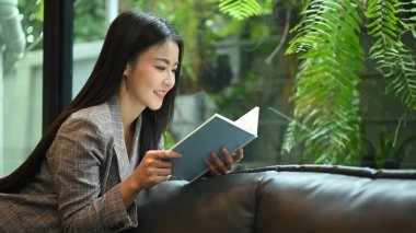 Elegant millennial business woman resting on couch in bright modern office and reading book.