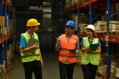 Mature male manager and workers in hardhats and reflective jackets checking quantity of storage product on shelf in a large warehouse.