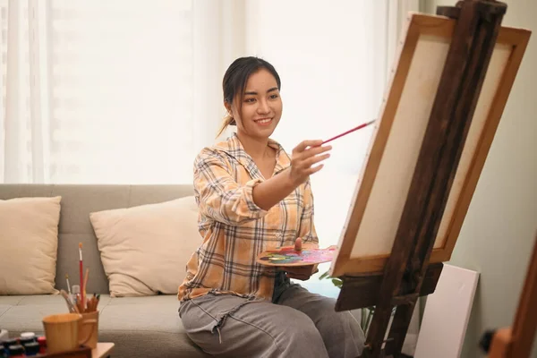 Young beautiful woman artist painting on canvas with oil colors in bright living room. Hobby, art and people concept.