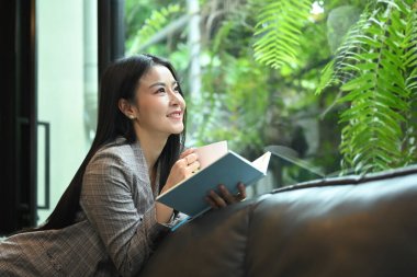 Beautiful millennial woman reading book while resting on couch at her office.