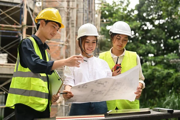 Team of civil engineers and real estate investor wearing safety helmets and vests examining plans at construction site.