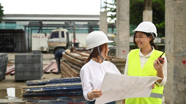 Smiling female real estate investor and civil engineers manager wearing safety helmets examining plans at construction site.