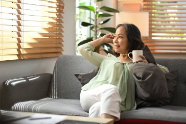 Retired woman resting on comfortable couch drinking hot coffee, enjoy stress free peaceful mood at the morning.