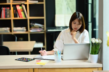 Focused young creative woman entrepreneur watching webinar or working on her laptop at home office.