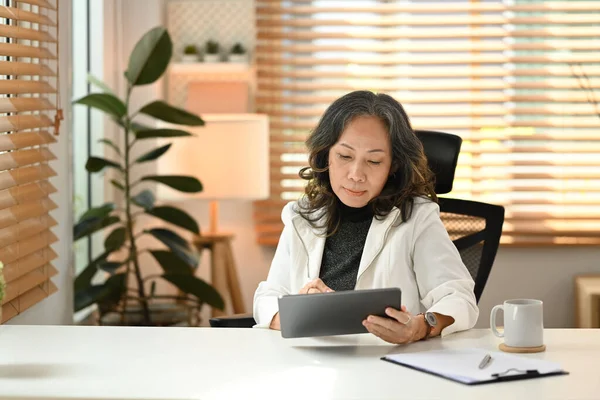 Attractive mature businesswoman checking email on digital tablet during working online from home.