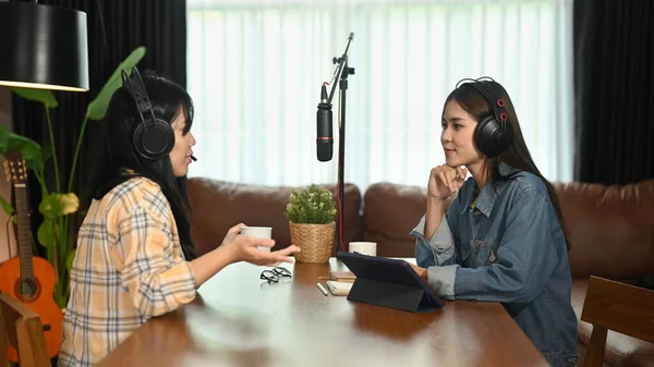 Two young female bloggers in headphones looking and talking each other while recording conversation, interview for their channel.