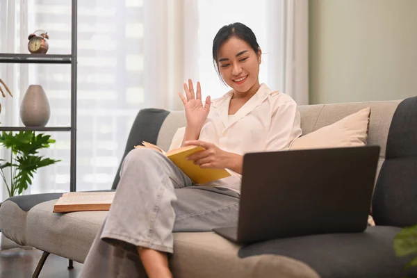 Happy millennial woman waving hands and making video call via laptop while resting on couch at home.