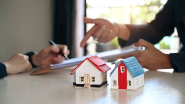 Two small house models on white table with costumer signing contract with agent in background. Real estate and Property purchase concept.