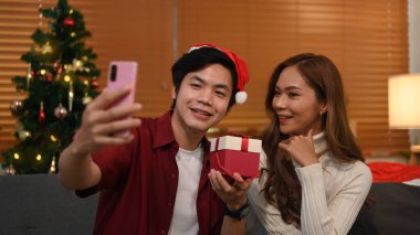 Happy young couple taking selfie, making video call on smart phone and celebrating Christmas holidays together at home.