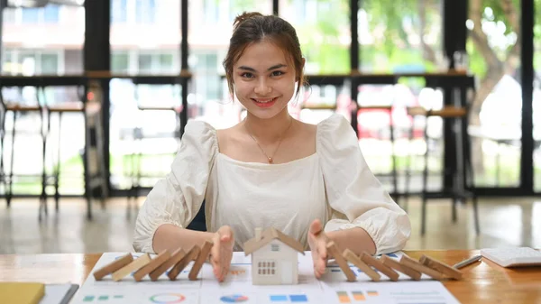 Smiling young woman protecting house model from falling wooden blocks with hands. Home insurance, risk and problem.