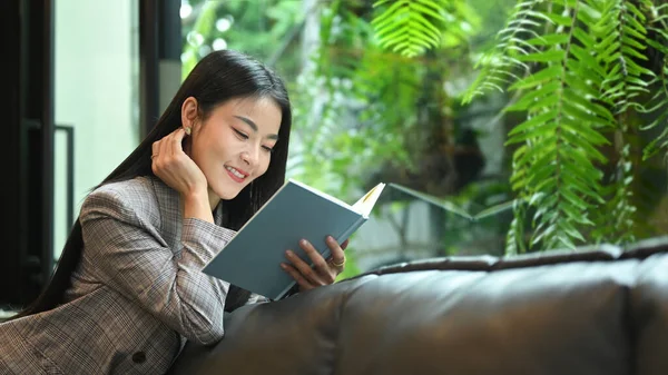 Attractive millennial woman reading book on couch in beautiful day. Leisure activity concept.