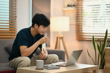 Asian man in casual clothes sitting in cozy living room and browsing internet with laptop computer.