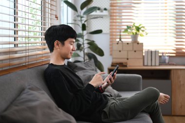 Satisfied man in warm sweater resting on couch and using mobile phone, enjoying chatting online with his friends.
