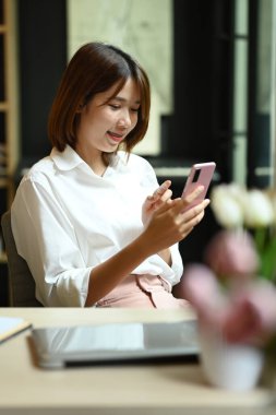 Charming young asian woman using smart phone sitting in bright modern corporate office.