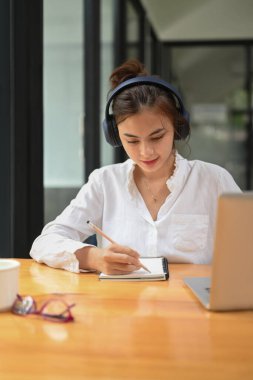 Concentrated female student wearing headphone, listening lecture during study online on laptop and making notes on notebook.