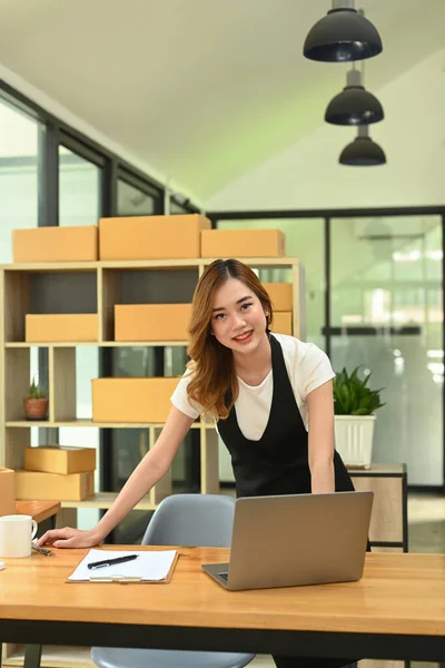 Happy young woman online seller standing front of laptop in home office and smiling to camera. Online selling, E-commerce, Online Shopping.