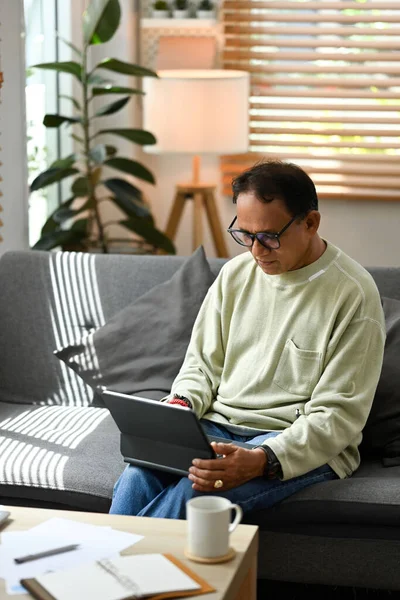 Happy mature man sitting on couch and checking email, reading online news on tablet. Elderly technology concept.