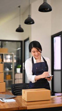 Önlüklü genç bir kadın, ürün satın alma siparişini kontrol ediyor, ev ofisinde çalışıyor. E-ticaret, çevrimiçi iş, çevrimiçi satış kavramı. 