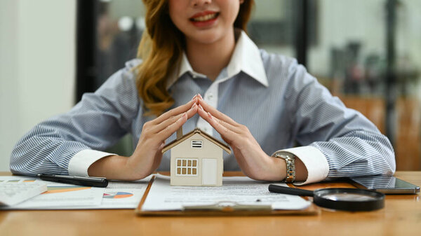 Улыбающаяся женщина защищает миниатюрный дом на деревянном столе. Страхование имущества и надежное финансирование спокойная концепция.