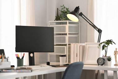 Rahat bir ofis. Boş ekranlı bilgisayar, kırtasiye, ev bitkisi ve lamba beyaz masada.