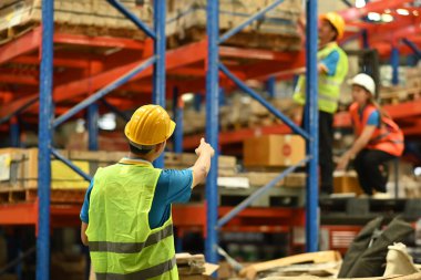 Erkek müdürün kasketli ve yansıtıcı ceketli arka planı üst rafı ve depodaki malları gösteriyor..