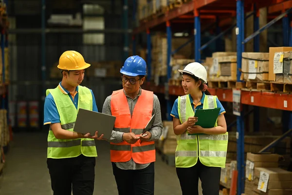 Middle aged manager and young workers inspecting stock and cardboard stock product on laptop computer in a large warehouse.