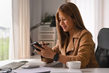 Gülümseyen yaratıcı kadın parlak ofiste oturuyor ve akıllı telefon kullanıyor..